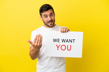 Wall Mural - Young handsome caucasian man isolated on yellow background holding We Want You board and doing coming gesture