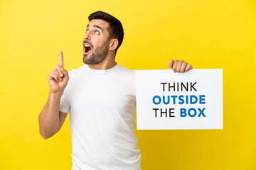 Wall Mural - Young handsome caucasian man isolated on yellow background holding a placard with text Think Outside The Box and thinking