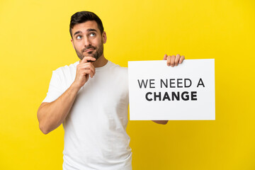 Wall Mural - Young handsome caucasian man isolated on yellow background holding a placard with text We Need a Change and thinking