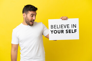 Wall Mural - Young handsome caucasian man isolated on yellow background holding a placard with text Believe In Your Self