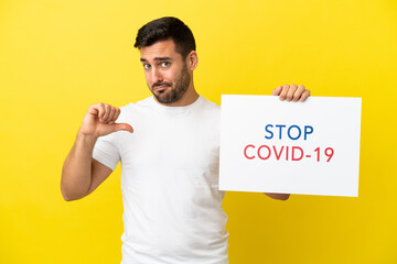 Wall Mural - Young handsome caucasian man isolated on yellow background holding a placard with text Stop Covid 19 with proud gesture