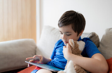 Wall Mural - Portrait young boy playing game on tablet sitting on sofa with light shining from window, Kid playing games on internet, Cinematic portrait Child doing homework online at home