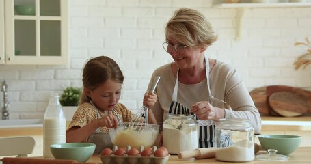 Wall Mural - Happy little 7s child girl enjoying cooking with joyful caring middle aged older grandmother in modern kitchen. Interested sincere different generations female family preparing weekend breakfast.