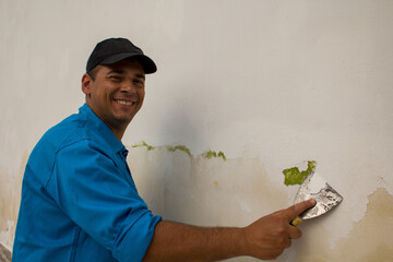 Wall Mural - Image of a smiling handyman who removes paint from a wall with a spatula to restore it from mold.