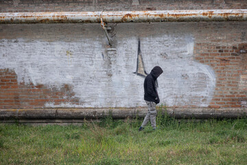 Boy with a Black Hoodie in his Head Walking on the Grass Next to a Brick City Wall