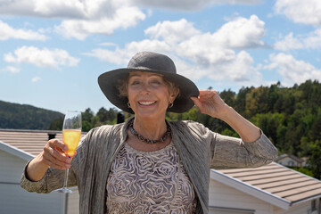 a woman of retirement age in excellent physical shape and with an open smile, dressed in a dress and a hat, holds a glass of champagne in her hand against a cloudy sky, the concept of an active lifest