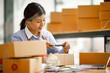 © David - Young Asian entrepreneur SME woman checking product on stock take note with a laptop. Small business owner working at home. Check address on the parcel SME online start up freelance concept.