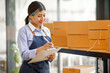 © David - Young Asian entrepreneur SME woman checking product on stock take note with a laptop. Small business owner working at home. Check address on the parcel SME online start up freelance concept.