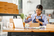 © David - Young Asian entrepreneur SME woman checking product on stock take note with a laptop. Small business owner working at home. Check address on the parcel SME online start up freelance concept.