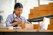 © David - Young Asian entrepreneur SME woman checking product on stock take note with a laptop. Small business owner working at home. Check address on the parcel SME online start up freelance concept.
