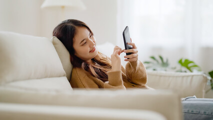 Happy young asian woman relax on comfortable couch at home texting messaging on smartphone, smiling girl use cellphone, browse wireless internet on gadget, shopping online from home