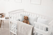 Baby crib with blanket, pillows and teddy bear near light wall