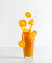 Glass With Splashing Of Orange Juice And Falling Orange Slices On Table At White Background. Healthy Refreshing Drink. Liquid Motion. Front View.