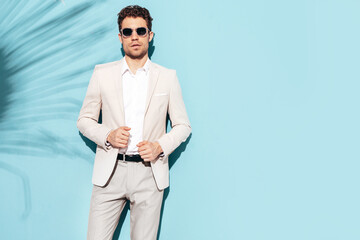 Portrait of handsome confident stylish hipster lambersexual model. Sexy modern man dressed in white elegant suit. Fashion male posing in studio near blue wall in sunglasses