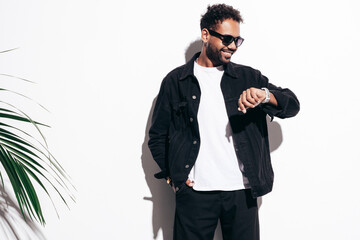 Handsome smiling hipster model. Sexy unshaven man dressed in black summer shirt and jeans clothes. Fashion male posing near white wall in studio at sunny day. In sunglasses. Looking at watch