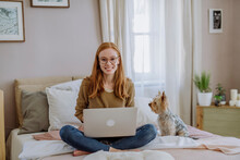 Smiling Woman Sitting With Laptop By Pet Dog On Bed At Home