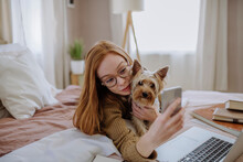 Smiling Woman Taking Selfie With Pet Dog Through Smart Phone Lying On Bed At Home