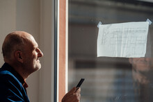 Businessman Holding Mobile Phone Analyzing Blueprint On Window In Office