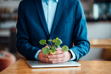 Freelancer Holding Plant Over Laptop At Table In Cafe