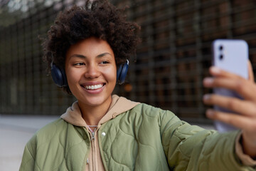 Wall Mural - positive curly teenage girl blogger listens music via headphones has online communication shoots vid