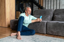 Mature Man Using Mobile Phone Sitting By Sofa At Home