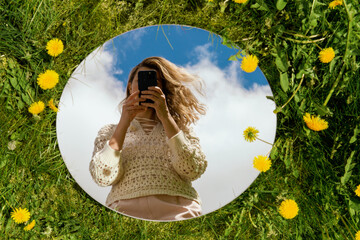 nature and people concept - woman with smartphone reflection in round mirror on summer field with dandelion flowers