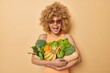 © wayhome.studio  - Positive beautiful woman embraces bunch of fresh green vegetables and fruits wears casual t shirt and sunglasses focused above with glad expression dressed in t shirt isolated over beige background