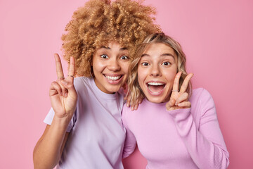 Wall Mural - Positive happy women have fun carefree mood make peace gesture or v sign have glad expressions stand closely dressed in casual clothes isolated over pink background express optimism and joy.