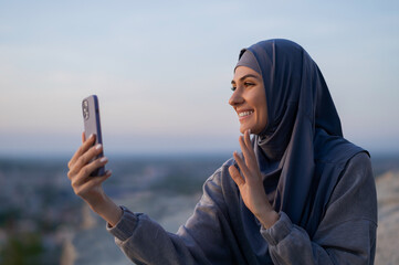 Wall Mural - Young beautiful girl in a hijab speaks by video link on the phone and shakes hands on a background of sunset and cityscape