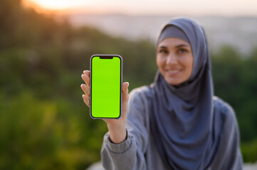 Wall Mural - Phone with green screen in the hands of a Muslim girl in a hijab on a background of sunset and landscape