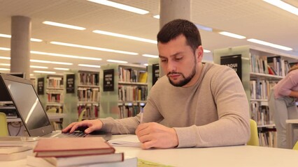 Canvas Print - Focused guy student preparing for an exam on a laptop in the university library, looking for sources of information in textbooks. High quality 4k footage