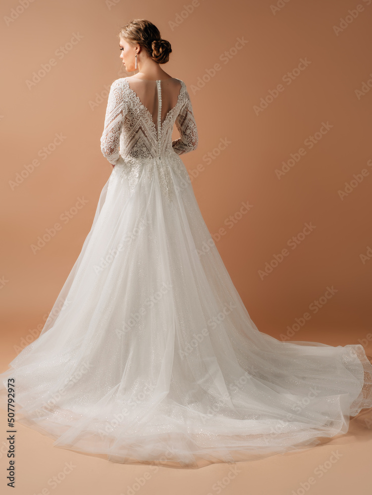 Photo Stock Luxury shiny lace wedding dress. Summer backless long sleeve  bridal gown with tulle skirt. Beautiful blonde bride posing in studio on  brown background. Natural looking makeup and hairstyle, rear view. |