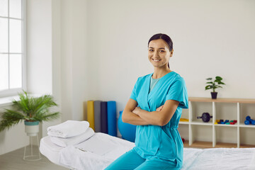 Wall Mural - Portrait of friendly confident professional female masseur or chiropractor in her medical office. Young woman in blue medical uniform sits on massage couch with folded arms and looks at camera.