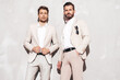 Portrait of two handsome confident stylish hipster lambersexual models. Sexy modern men dressed in white same elegant suit. Fashion male posing in studio near grey wall