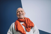 Portrait of cheerful senior man wearing orange scarf standing against blue and white wall