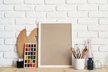 Photo Frame As Easel With Artist's Tools On Wooden Table Against White Brick Wall