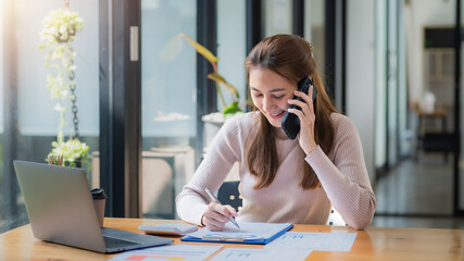 Smiling young Asian woman talking on the phone and working at home.