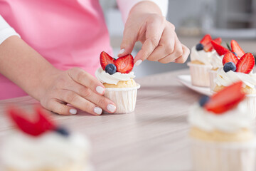 Pastry chef confectioner young caucasian woman in the kitchen. She is decorating a cupcake with with strawberries and blueberries