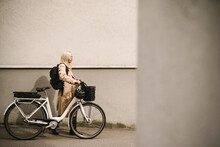 Side View Of Businesswoman Wheeling With Electric Bicycle By Wall