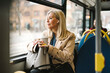 © Maskot - Businesswoman looking through window holding disposable cup while commuting in bus
