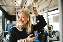 Happy Businesswoman Talking On Smart Phone Holding Grab Handle While Commuting Through Bus