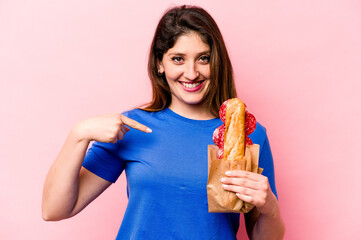 Young caucasian woman eating a sandwich isolated on pink background person pointing by hand to a shirt copy space, proud and confident