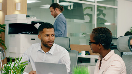 Two african workers are at the modern office and they are sitting at their desks smiling and agreeing with one another, in the back a man and woman are working at the printer