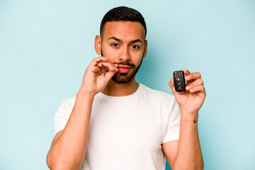 Wall Mural - Young hispanic man holding car keys isolated on blue background with fingers on lips keeping a secret.