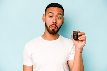 Wall Mural - Young hispanic man holding car keys isolated on blue background shrugs shoulders and open eyes confused.