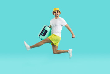 Joyfully excited guy in summer clothes runs and jumps with suitcase on light blue background. Young man in shorts, panama and t-shirt in hurry summer vacation with crazy funny facial expression.