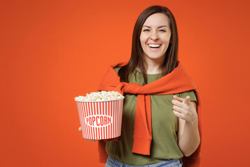 Wall Mural - Young smiling laughing fun happy woman in khaki t-shirt tied sweater on shoulders point index finger camera on you hold bucket of popcorn isolated on plain orange background. People lifestyle concept.