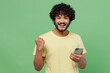 © ViDi Studio - Young happy Indian man 20s in basic yellow t-shirt hold in hand use mobile cell phone chatting online do winner gesture isolated on plain pastel light green background studio People lifestyle concept.