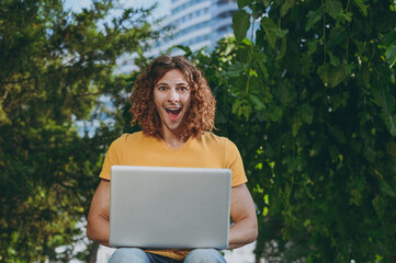 Surprised amazed excited fun cool young man 20s wearing yellow t-shirt holding use work on laptop pc computer rest relax in spring green city park outdoors on nature. Urban lifestyle leisure concept