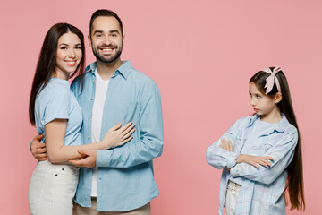 Young happy mom dad with sad jelaous child kid daughter teen girl wearing blue clothes look at hugging parents isolated on plain pastel light pink background. Family day parenthood childhood concept.